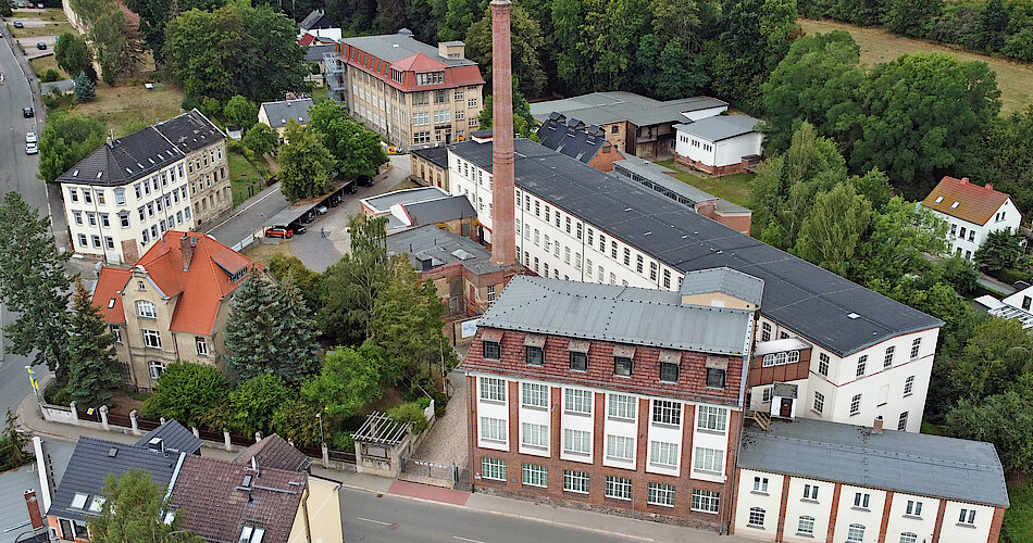 Das Foto zeigt das gesamte Gelände der Tuchfabrik Gebr. Pfau. Es ist ein Luftbild, welches mit einer Drohne aufgenommen wurde. Im Mittelpunkt ist der Schornstein zu sehen, umgeben von den verschiedenen Gebäuden der Fabrik.