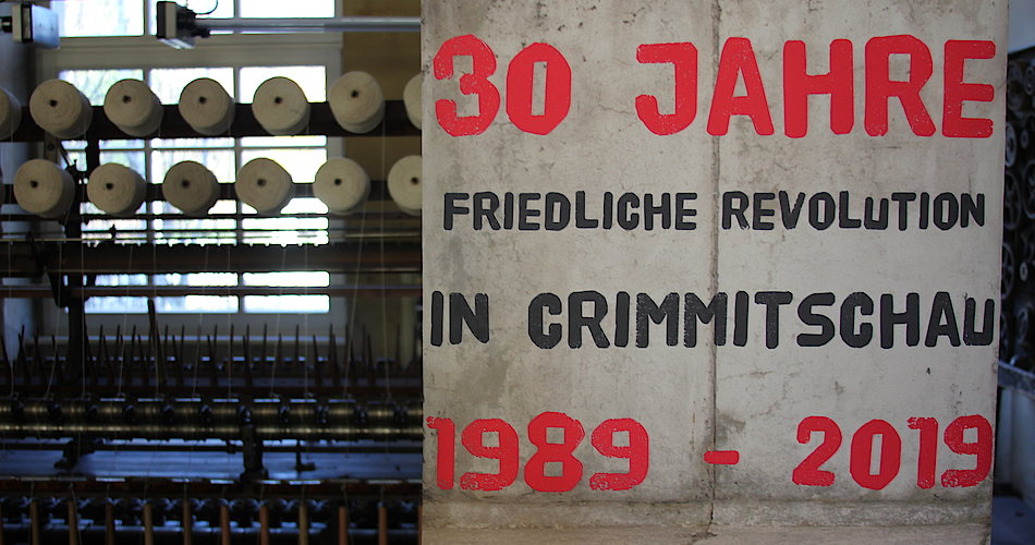 Im Vordergrund befindet sich das Plakat zur Ausstellung mit dem Titel "30 Jahre friedliche Revolution in Crimmitschau 1989 - 2019". Im Hintergrund ist ein Teil des Ausstellungsraumes zu sehen mit einer der Zwirnmaschinen der Tuchfabrik.