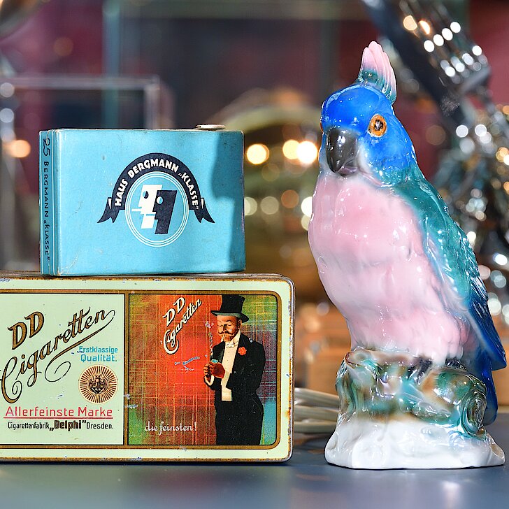 Das Foto zeigt einen Rauchverzehrer in Form eines Papageien und zwei Zigarettenschachteln der Marken "Haus Bergmann" und "DD Cigaretten".