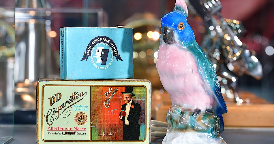 Das Foto zeigt einen Rauchverzehrer in Form eines Papageien und zwei Zigarettenschachteln der Marken "Haus Bergmann" und "DD Cigaretten".